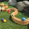 Alder-wood curvy tracks set on green artificial grass with colorful balls (orange, pink, yellow, green, blue, and purple) placed throughout the tracks. Large rocks and a Grimm's Large Natural Rainbow Stacker decorate the background, adding a natural element to the playful setup. The materials are all non-toxic for safe play.