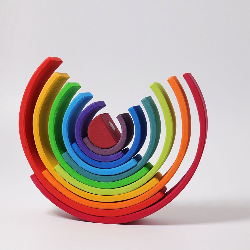 Grimm's Large Wooden Rainbow is a versatile, colorful wooden stacking toy arranged in an open semi-circle. Made from non-toxic wood, the pieces are painted in varying shades of the rainbow, starting with red on the outermost edges and progressing to purple in the center. Each curved piece fits neatly within one another.