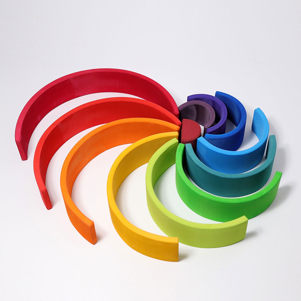 A colorful arrangement of semi-circular wooden blocks, each in a different hue, forms a circular pattern on a white surface. This versatile toy, known as Grimm's Large Wooden Rainbow, features shades of red, orange, yellow, green, blue and purple. The blocks are made from non-toxic wood for safe play.