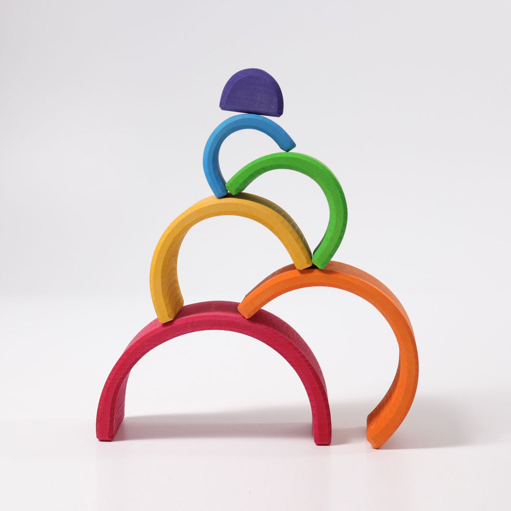 A vibrant display featuring Grimm's Wooden Rainbow, showcasing an array of non-toxic wooden arches stacked in a harmonious rainbow arrangement against a white background. Starting from the bottom, the colors gracefully ascend from red to orange, yellow, green, and blue, crowned with a blue semicircle at the apex. This versatile toy invites boundless imaginative play.