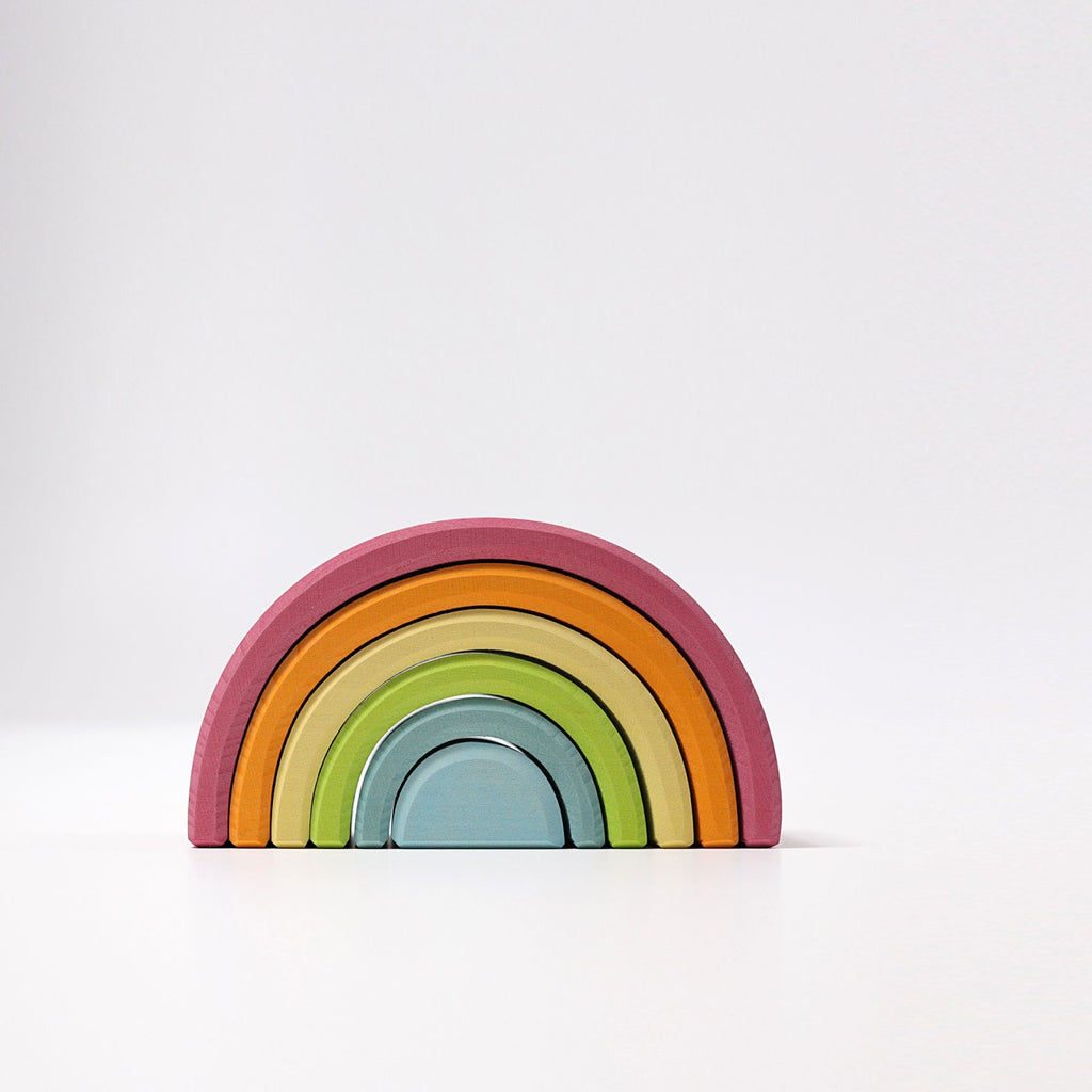 A Grimm's Rainbow Pastel Stacker arranged in the shape of a rainbow tunnel. The set includes several arc-shaped pieces painted in a gradient of colors: red, orange, yellow, green, and blue, from top to bottom. Perfect for toddlers to stack and made with non-toxic materials. The background is plain white.