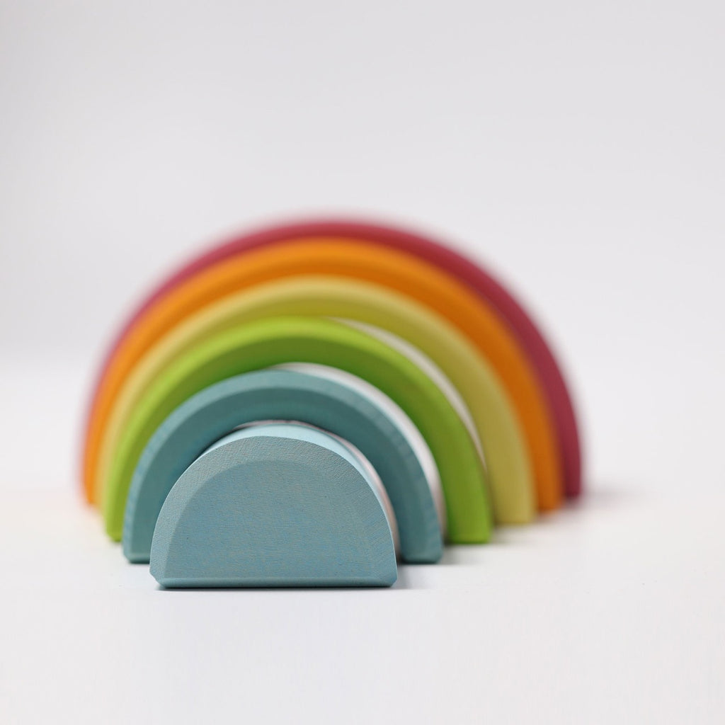 A set of Grimm's Rainbow Pastel Stacker in non-toxic rainbow colors arranged in a row on a white background. The blocks are different sizes, with the smallest in blue in the foreground and the largest in red at the back, creating a rainbow tunnel effect perfect for toddlers to stack.