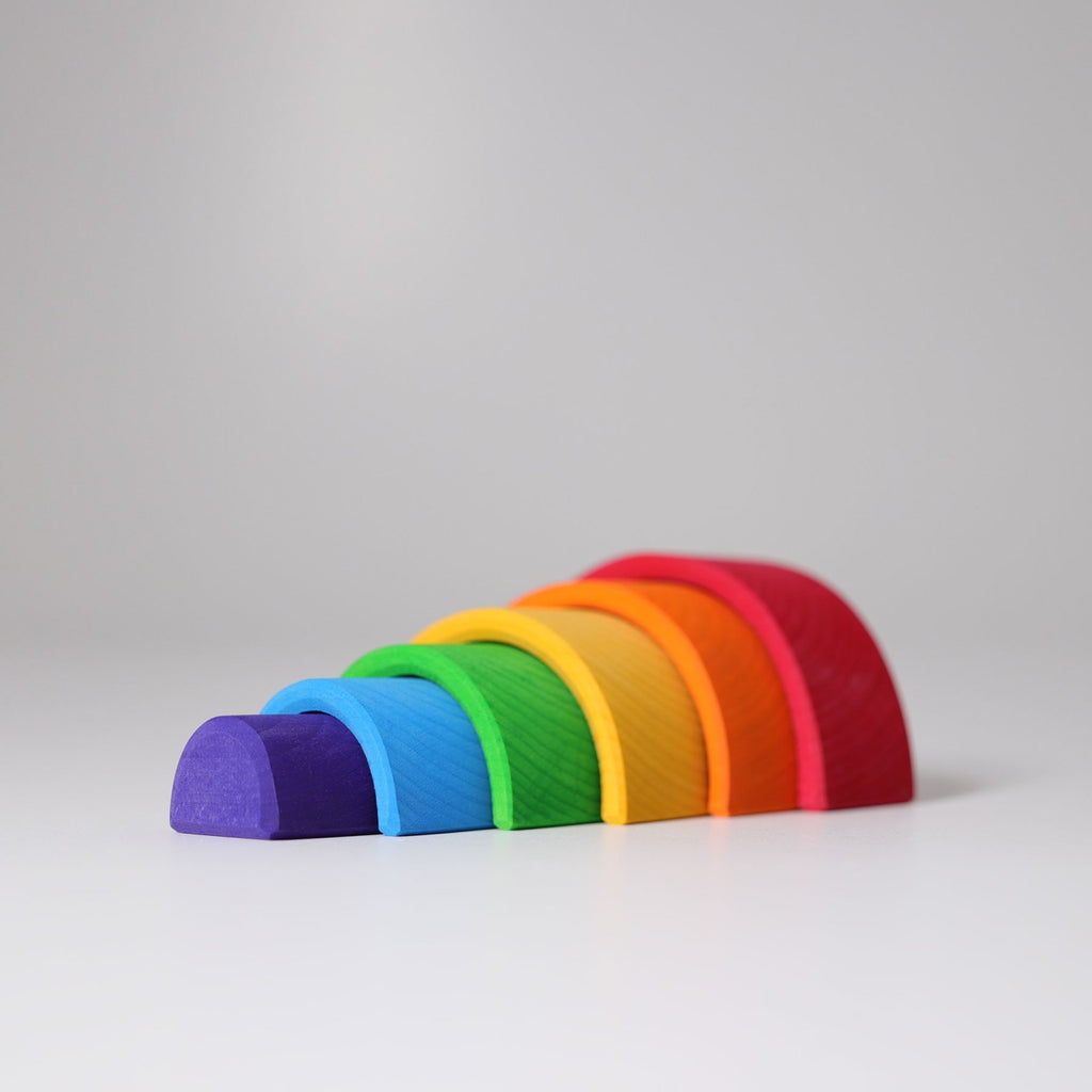 A minimalist photo of a Grimm's Small Wooden Rainbow, also known as a rainbow tunnel, arranged on a white surface. The versatile toy consists of seven semi-circular arches in descending size order from red, orange, yellow, green, blue, purple to dark purple, creating a gradient effect.