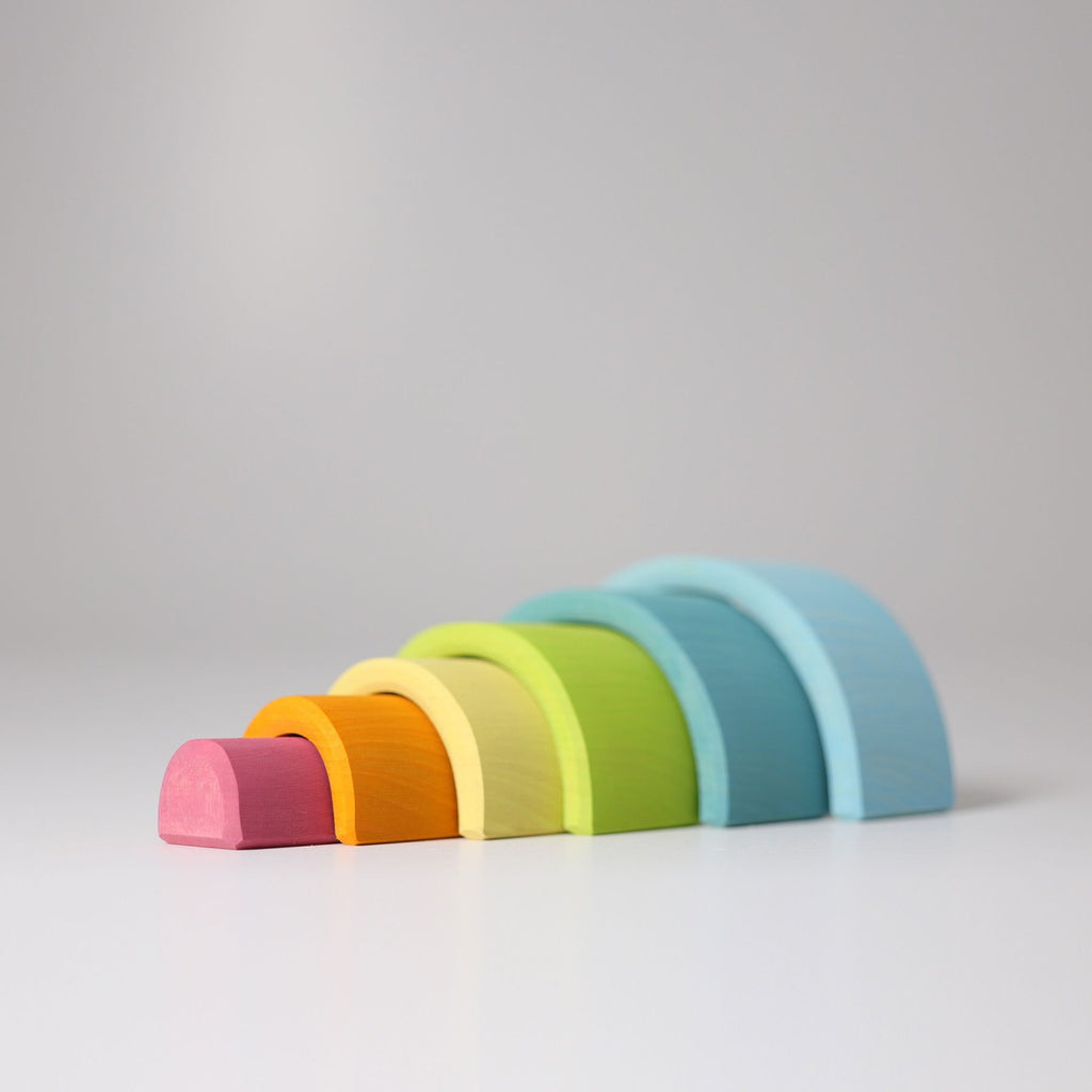 A minimalist photo displays Grimm's Small Wooden Rainbow, Pastel arranged in a gradual size order. The blocks, resembling a versatile toy, are colored pink, orange, yellow, green, blue, and light blue against a plain light grey background.