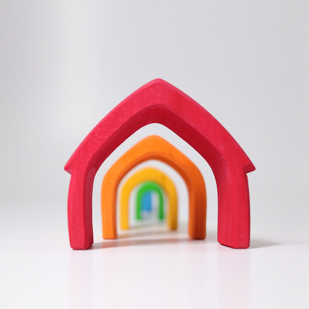 A set of colorful, arch-shaped blocks, perfect for toddlers, are arranged in descending order of size from front to back, creating a tunnel effect. The Grimm's Colorful House Stacker features arches in red, orange, yellow, green, and blue on a white surface against a plain white background.