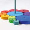 A variety of colorful wooden discs, available in blue, green, yellow, orange, and purple and of different sizes, are neatly arranged on a flat surface with a vertical rod at the center. The arrangement forms a Grimm's Stacking Tower in progress. This engaging toy enhances eye-hand coordination and fine motor skills. The background is white.