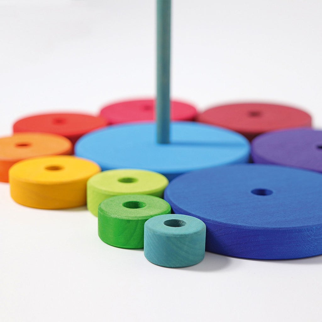 A variety of colorful wooden discs, available in blue, green, yellow, orange, and purple and of different sizes, are neatly arranged on a flat surface with a vertical rod at the center. The arrangement forms a Grimm's Stacking Tower in progress. This engaging toy enhances eye-hand coordination and fine motor skills. The background is white.