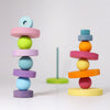 A minimalist image showcasing stacks of colorful, smooth, wooden discs and spheres balanced one on top of the other in two piles. A third stack's base and supporting stick for this Pastel Stacking Tower are visible in the background on a white surface. The discs and spheres in varying pastel shades foster fine motor skills.