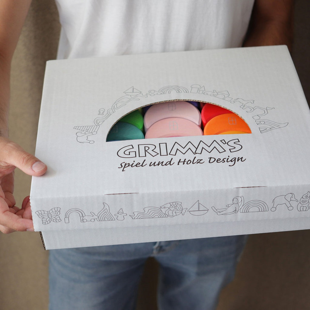 A person holds a white box labeled "Grimm's Moon Houses," displaying colorful wooden toys inside. Perfect for developing fine motor skills, the box is adorned with line drawings of playful objects that encourage children to sort and stack.