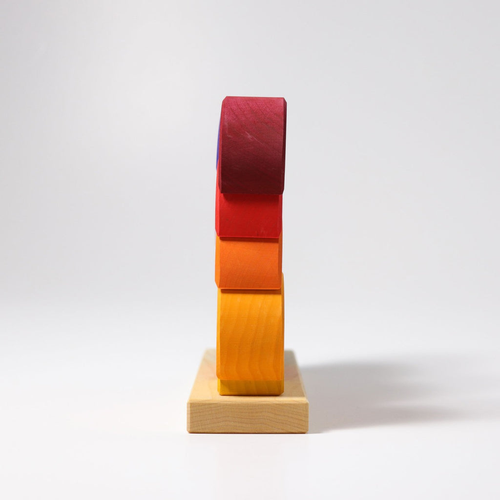 The Grimm's Moon Houses is a minimalist sculpture showcasing stacked wooden blocks in varying colors—purple, red, orange, and yellow—mounted on a square wooden base. The elegant arrangement encourages engagement with fine motor skills by creating a curving effect against a plain white background.