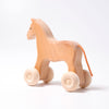 A Grimm's Wooden Horse Willy Push Toy with a smooth, natural wood finish. The horse has simple, carved details, a small brown string tail, and four wooden wheels attached to its legs for easy rolling. Made in Germany with non-toxic materials, the toy is pictured against a plain white background.