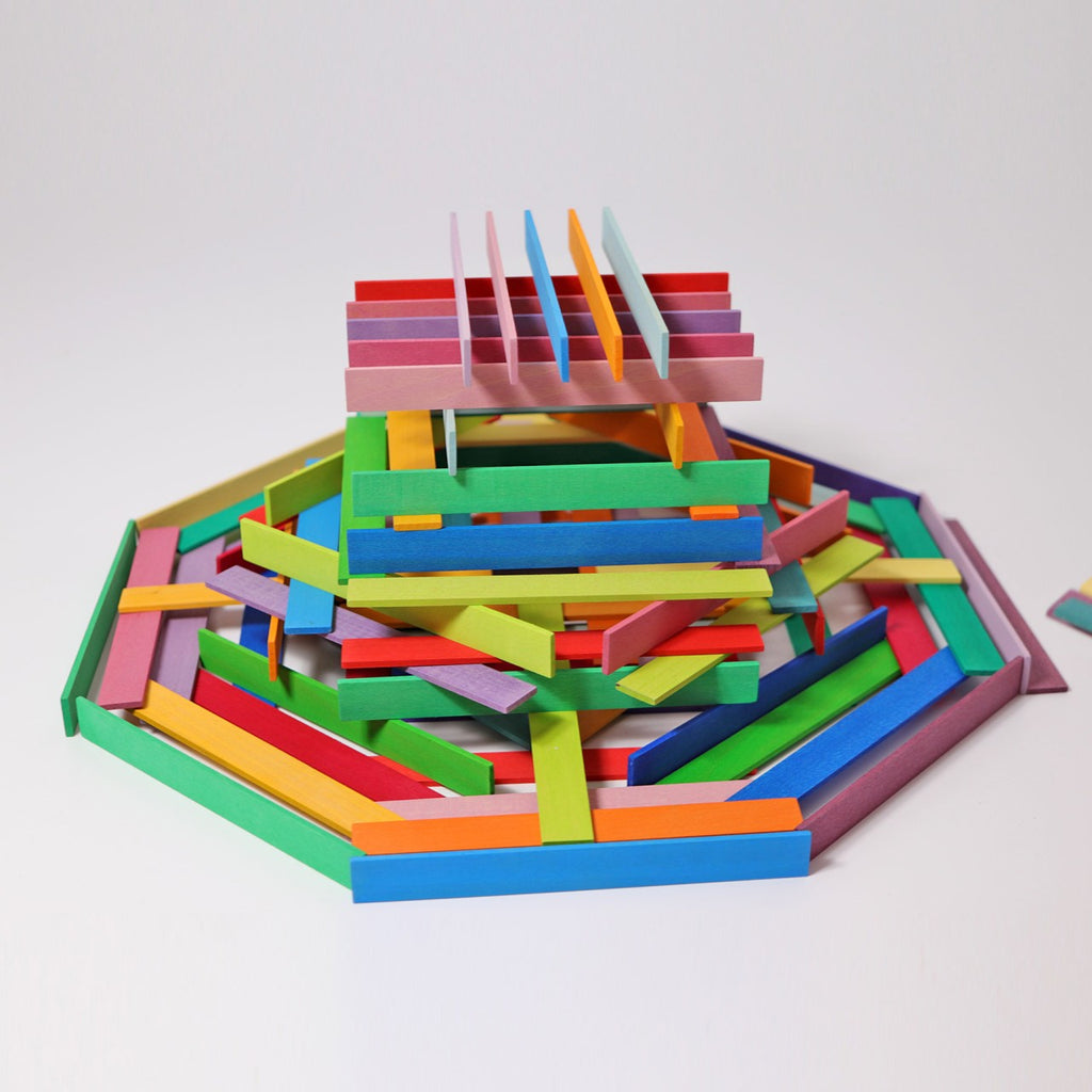 A colorful, intricate children's building toy made of interlocking, rectangular wooden blocks is displayed on a white background. The blocks are arranged in various layers and orientations, creating a vibrant, geometric design with red, pink, yellow, blue, green, and purple pieces that encourage fine motor skills development. Grimm's Leonardo da Vinci Building Sticks are perfect for inspiring creativity and educational play among young children.