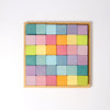 A neatly arranged set of Grimm's Pastel Mosaic Building Blocks in a wooden tray. The blocks, in shades of blue, pink, orange, yellow, green, and purple, form a grid pattern of five rows and five columns on a white background. This engaging toy helps children develop fine motor skills.