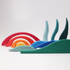 A collection of colorful, curved wooden pieces in the shapes of waves and arches, resembling landscape building blocks, are arranged artistically against a plain white background. The vibrant colors include red, orange, yellow, light blue, blue, and green. This is the Grimm's Creative Puzzle Landscape Block Set.