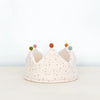 A soft, fabric Confetti Reversible Birthday Crown with a white base and small, multicolored dots. This handmade crown features five triangular points, each topped with a different colored button: green, orange, blue, and yellow. Made of organic cotton, it boasts a clean, light gray background.