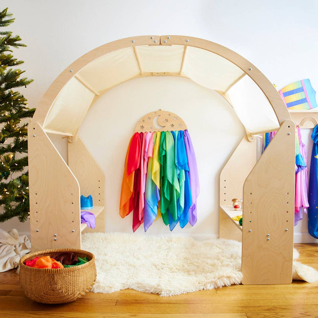 A wooden arch stands over a white furry rug, with colorful fabric draped from a crescent moon at the back. To the left, you'll find a woven basket filled with toys and vibrant Sarah's Silk Giant Lambswool Playsilks in winter colorways, while a small decorated Christmas tree is partially visible beside it.