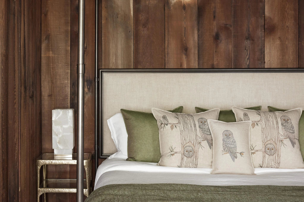 A cozy bedroom features a bed with a beige headboard against a wooden wall. The bed is adorned with 100% linen fabric in green and white, and several pillows including the Coral & Tusk Owl Family Tree Pillow. A small metallic nightstand with a white lamp is next to the bed.