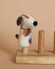 A small needle felted Calico Dog Finger Puppet with white, brown, and orange patches stands on a wooden base with two cylindrical posts. The dog has a black nose and wears a tiny bell around its neck. Handmade in Nepal, the background is a solid beige color.