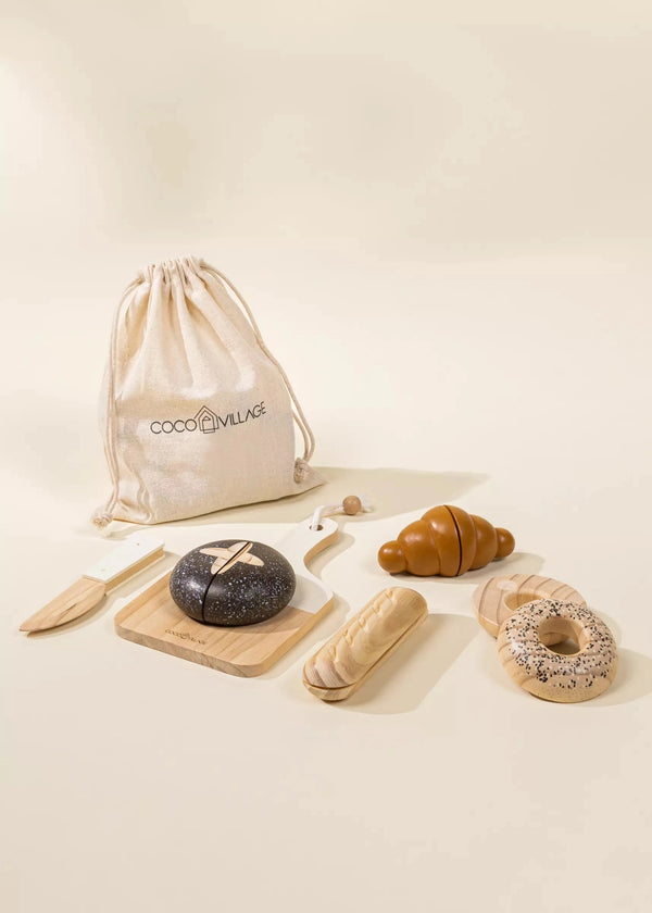 The Wooden Bakery Set features an artfully arranged selection of wooden toy bakery items, including various types of bread like a croissant, a baguette, a donut, and a black round loaf displayed on a wooden cutting board. Also included are a wooden knife and a cloth bag labeled "Coco Village." The set is showcased against a light beige backdrop, perfect for promoting healthy eating habits in children's playtime.