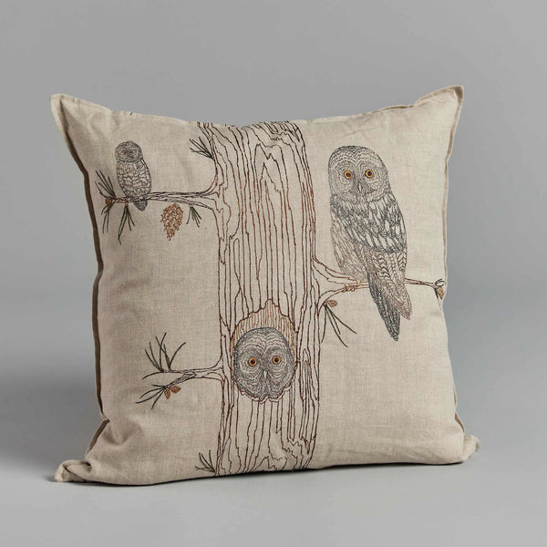 The Coral & Tusk Owl Family Tree Pillow, made from 100% linen fabric, showcases a beige cushion illustrating a tree trunk decorated with three embroidered owls: one sits on a branch at the top right, another peeks out from a hole in the trunk, and a smaller owl perches on a branch at the top left. The background is plain gray.