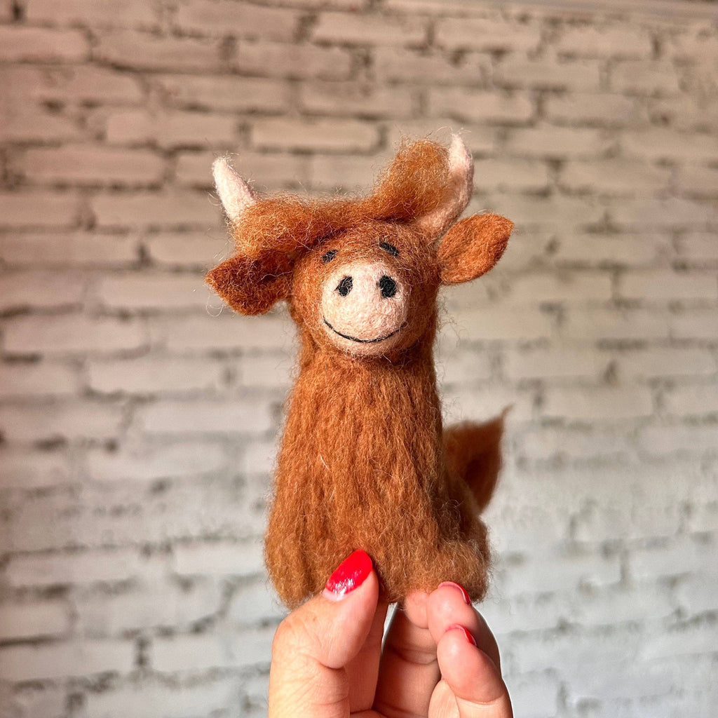 A Highland Cow Finger Puppet, crafted from brown felted wool with a smiling face, is displayed against a white brick wall backdrop. This small plush features large ears and horns, showcasing the craftsmanship of skilled Nepalese artisans. A hand with red-painted nails holds the adorable puppet.