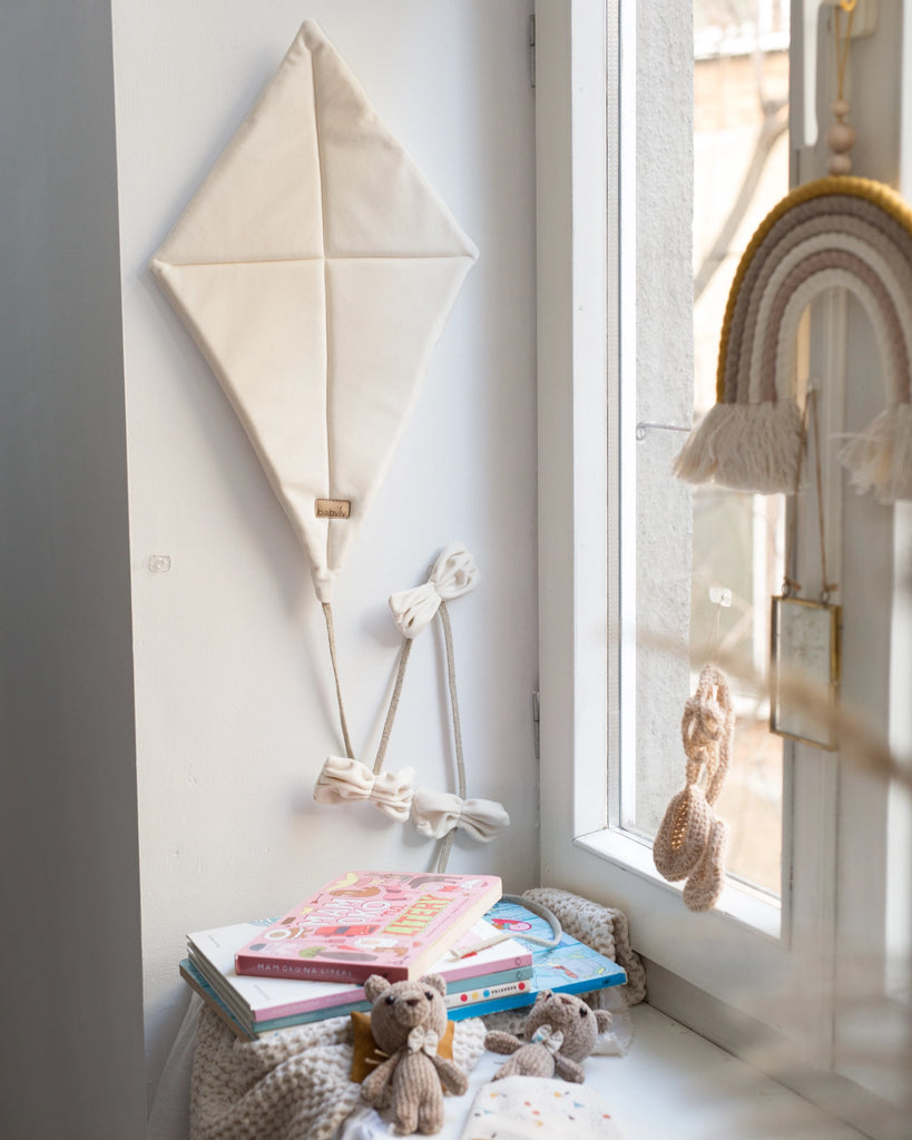 A cozy reading nook with a stack of children's books and plush toys on a cushioned seat by the window. A Velvet Kite Wall Decor - Cream and rainbow hanger adorn the wall, adding a whimsical touch to the calm ambiance. Sunlight filters through the window, creating an inviting atmosphere for any children's room decoration.