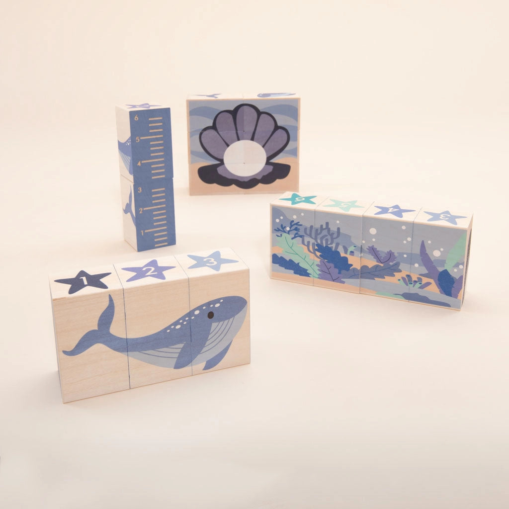 A set of Ocean Blocks crafted from sustainable basswood with ocean-themed illustrations is arranged on a light surface. The blocks display images of a blue whale, a shell, and an underwater coral reef scene. Some blocks have numbers and stars on them.