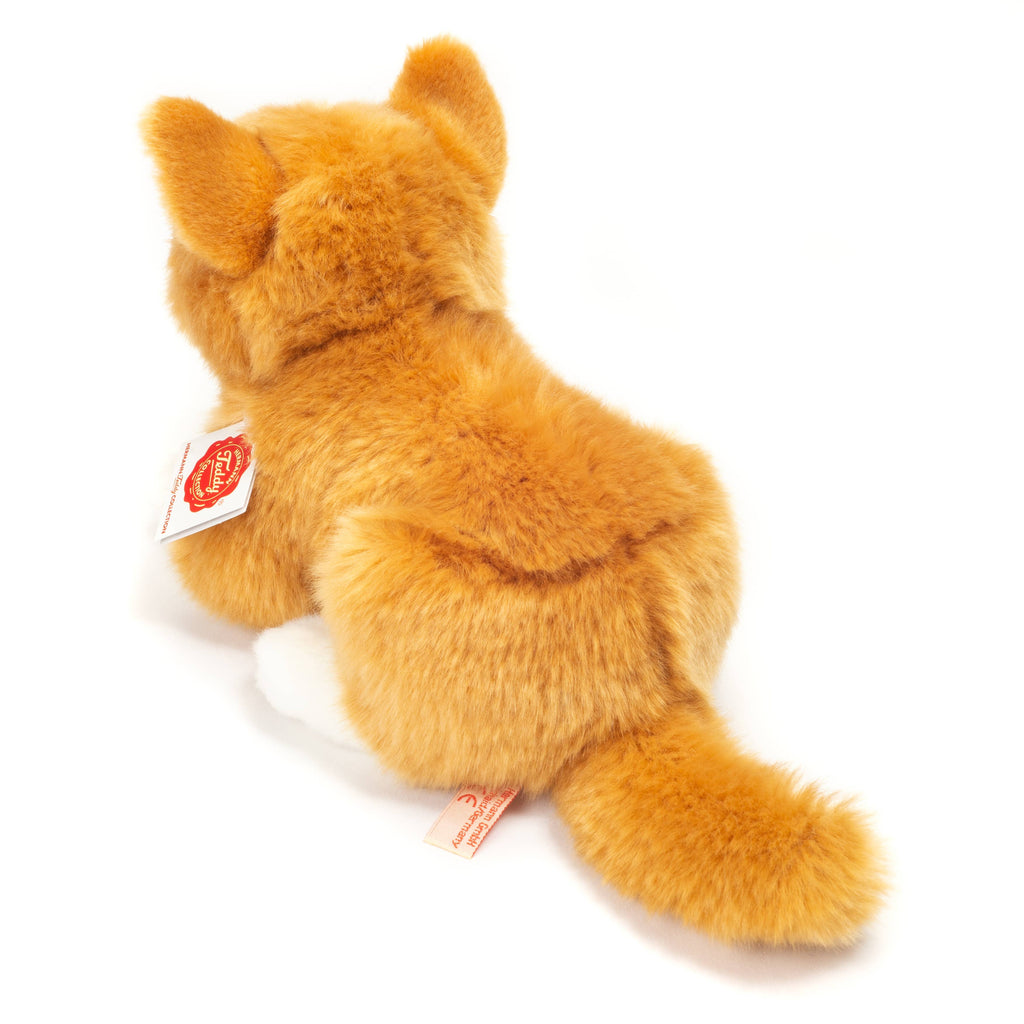 A fuzzy plush toy of a cat lying on its belly, facing away. The toy, part of the high-quality Hermann Teddy Collection, has soft orange fur with darker markings and white paws. Two tags are attached to its side and tail, with visible text and logos.

