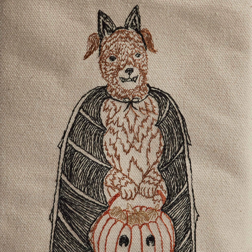 The Coral & Tusk Halloween Costumes Bag features an embroidered image of a dog standing on its hind legs, dressed as a bat with ears and wings, holding a Trick-or-treat bag. With its mischievous smile, this charming dog is perfect for adding to the fun among other costumed critters. The fabric background is beige.