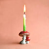 A lit green candle mounted on a hand-painted ceramic holder shaped like red and white spotted mushrooms, against a soft peach background.