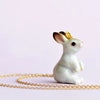 The White Rabbit Necklace features a delicate, hand-painted porcelain hare figurine adorned with a small 24k gold plated ring on its back. A charming thin gold chain partially coils around the base of the white rabbit, set against a soft, light pink background.