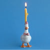 A whimsical Pink Goose Cake Topper wearing a pink bow tie and a polka dot party hat, with a lit yellow candle affixed to its back. The background is solid light blue.