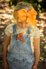 A young girl with blonde hair, wearing a large orange bow on her head, looks down at a stuffed animal in the front pocket of her denim overalls. She is also wearing a white shirt with orange polka dots and holds the Bubba Rooster Rattle. The background is blurred with green foliage.