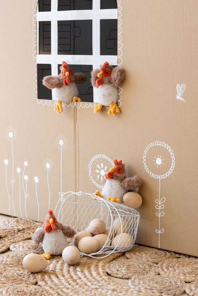 Three Bubba Rooster Rattles are arranged around a cardboard backdrop with a window. Two roosters are perched near the window, while one sits next to a wire basket filled with wooden eggs. The backdrop, reminiscent of action-reaction learning, is decorated with white flower illustrations and a butterfly drawing.