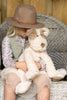 A child in a floral shirt and brown hat sits on a gray woven chair, holding their cherished Buddy Dog Stuffed Animal, creating a cozy, rustic scene.