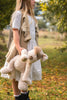 A young girl in a white dress and beige vest stands on a grassy path, holding her Buddy Dog Stuffed Animal. The scene is outdoors with green-leaved trees and a blurred landscape, evoking tranquility.