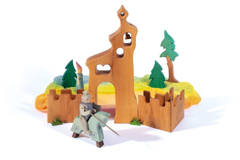 A handcrafted wooden toy set featuring a knight on horseback, a castle with walls, towers, green trees, and multicolored fabric representing the landscape. Made from sustainably sourced materials, the knight holds a lance and is dressed in a suit of armor, perfect for imaginative play with a medieval theme. The centerpiece of this set is the Ostheimer Bell Tower.