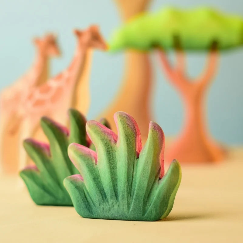 A close-up of the vibrant toy foliage, part of the Bumbu Wooden Bushes Set, with a blurred backdrop showcasing toy giraffes and a tree. The foliage pieces are predominantly green with accents of pink, meticulously crafted from sustainably sourced wood, and the set features toys that are artistically designed in soft, pastel hues.