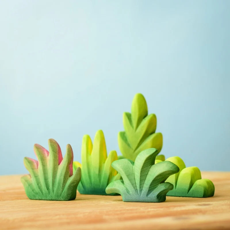 Colorful, abstract wooden toys resembling various types of green foliage are arranged on a wooden surface against a light blue background. This Bumbu Wooden Bushes Set, Montessori & Waldorf inspired, features different shades of green with hints of yellow and pink on some pieces.