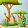 A colorful, whimsical scene featuring the Bumbu Wooden Bushes Set with eco-friendly wooden toy giraffes and lush green plants beneath large, stylized trees. The toys have a smooth, hand-painted appearance, creating a playful and imaginative forest environment against a light blue background.