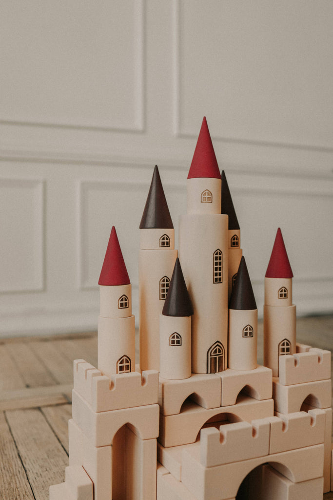 The Great Kingdom Building Set, featuring several tall towers with red and brown conical roofs, is set against a light-colored paneled wall. Crafted with smooth edges and non-toxic paint, the structure rests on a wooden floor, creating a whimsical and playful atmosphere.