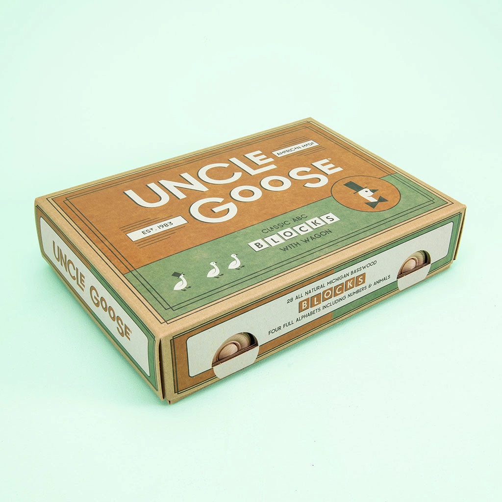 A cardboard box with "Uncle Goose Classic ABC Blocks with Wagon" written on it contains handmade alphabet blocks made from Michigan basswood. The design features illustrations of three geese and wooden wheels, evoking a charming basswood wagon, all set against a light green background.