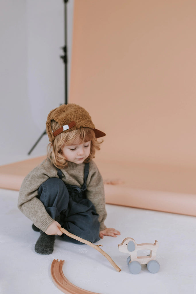 A young child with long hair, wearing a fuzzy brown hat with ear flaps, a gray sweater, and dark overalls, is sitting on the floor playing with a Wooden Puppy Push Toy. The background features a peach-colored backdrop. This activity is perfect for developing fine motor skills.