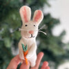 A hand holding a Bunny Finger Puppet, crafted with white needle felt and featuring pink inner ears and nose. The bunny, adorned with black eyes and whiskers, clutches an orange felt carrot topped with green foliage. The background is a blurred green setting, suggesting nature or foliage. Handmade in Nepal.