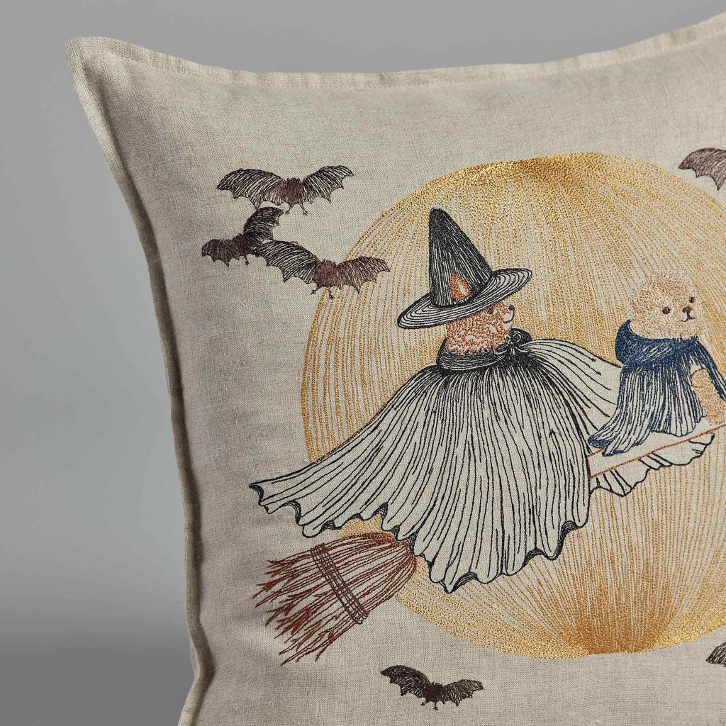 The Coral & Tusk Supermoon Spell Pillow showcases an illustration of a small figure in a flowing cape and pointy hat riding a broomstick, holding a Wizard Puppy with a scarf, set against the backdrop of a glowing yellow moon and flying bats.