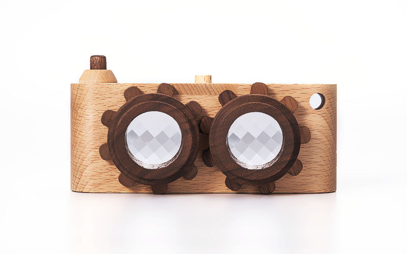 A Twinkle Kaleidoscopic Camera featuring two large circular kaleidoscopic lenses with gear-like edges, set against a white background. The body of the camera is made of light-colored wood with a small protrusion on the top left, mimicking a viewfinder in a retro stereo camera style.