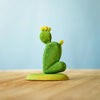 A small, green cactus sculpture with yellow dots from the Bumbu Wooden Cacti Set sits on a light wooden surface. The cactus features two arms, one raised higher than the other as if in a gesture of greeting. It rests against a soft blue gradient background that exudes serenity and minimalism.