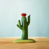 A small, handcrafted model of a green cactus with tiny white spots sits on a wooden surface against a light blue background. The cactus, part of the Bumbu Wooden Cacti Set, features two upward arms and a red flower blooming on top.