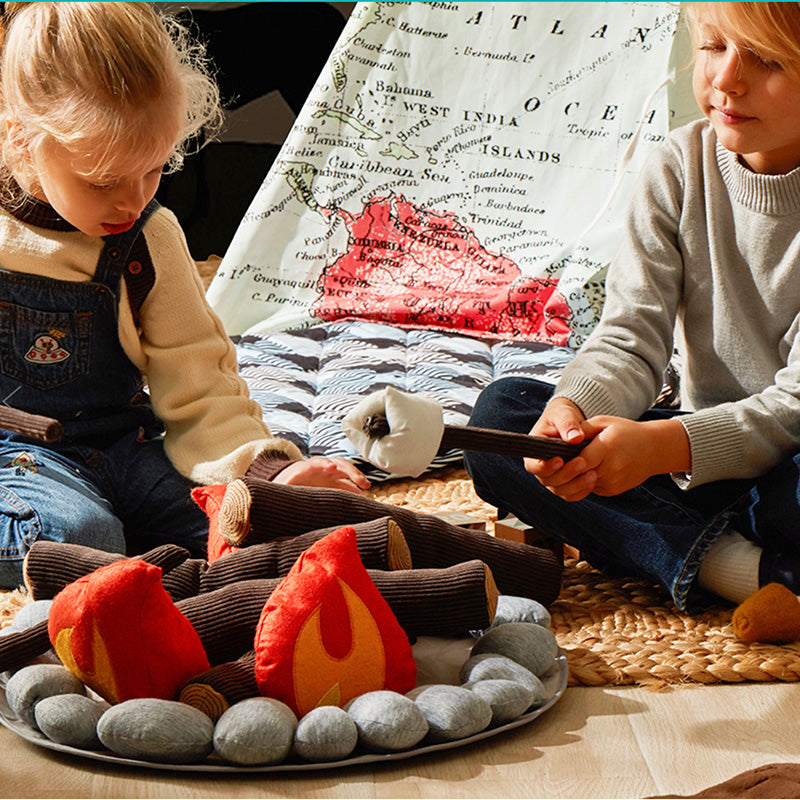 Two children are sitting on the floor playing with a Campout Campfire & S'mores. The campfire, made of faux felt logs and fabric flames, is surrounded by soft, gray stones. They're making pretend s’mores over the fire. Behind them, there's a tent featuring a world map design. The scene is cozy and playful.