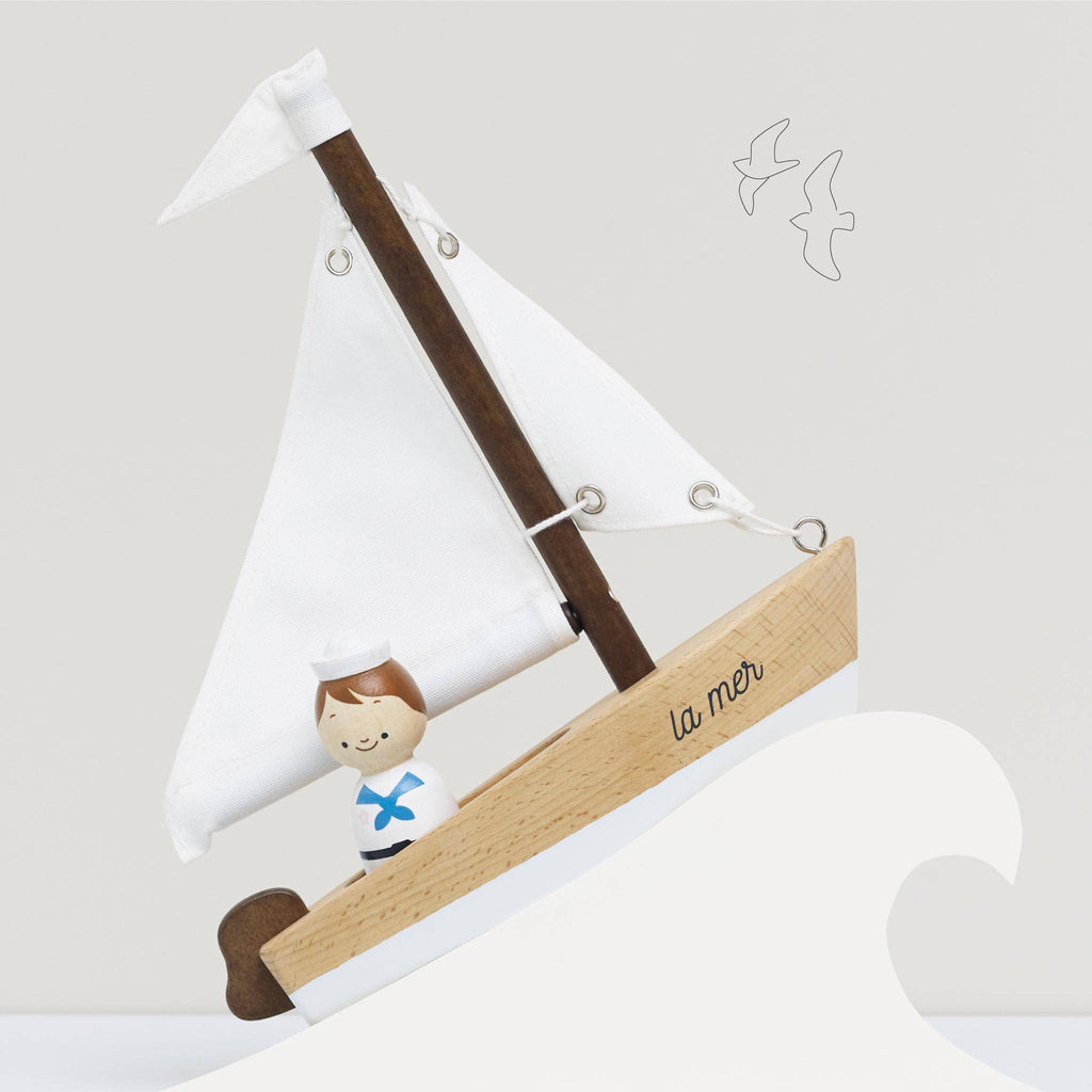 The Wooden Sailing Boat & Captain features a handcrafted design with a smiling sailor gliding on stylized waves. Its white sail is adorned with a blue cross, while "la mer" decorates the side, enhancing cognitive skills. The scene is completed by two birds drawn in the sky.